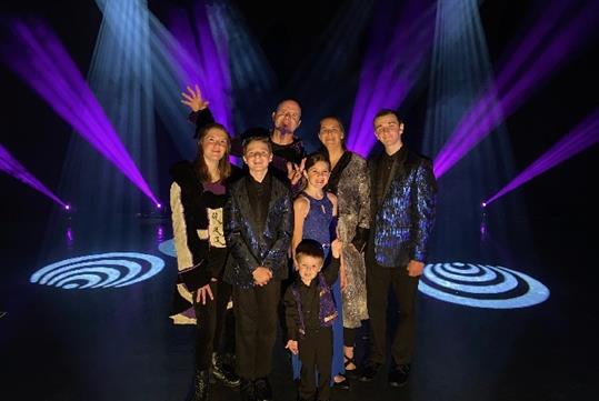 Phil Dalton and the rest of the Dalton family in costume, standing together on stage, are illuminated by white and purple lights.