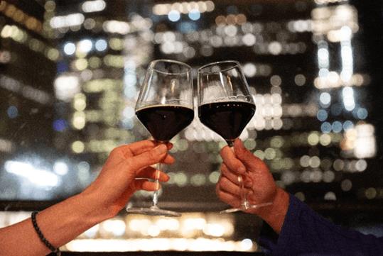 People cheering their glasses of red wine together with blurred city lights at night in the background.