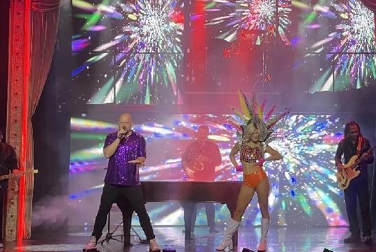 Kyle Martin performing on stage in a purple sequin shirt with a dancer next to him and a band in the background.