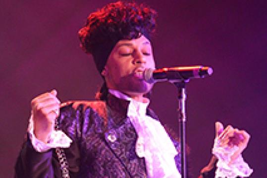 A Prince impersonator singing into a microphone wearing a purple jacket with a dark purple light shining on the stage.