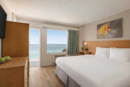 A modern hotel room with a white king bed, a dresser, art on the wall, a large window and an open door showing the ocean.