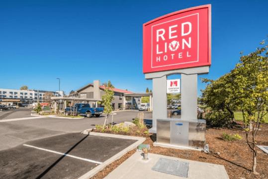 The red sign for the Red Lion Hotel in their parking lot with the hotel in the background on a sunny day in Portland.
