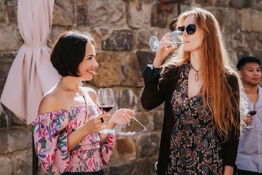 Two women standing in front of a stone wall drinking wine and smiling on a sunny day on the Redwoods and Wine Country Tour.