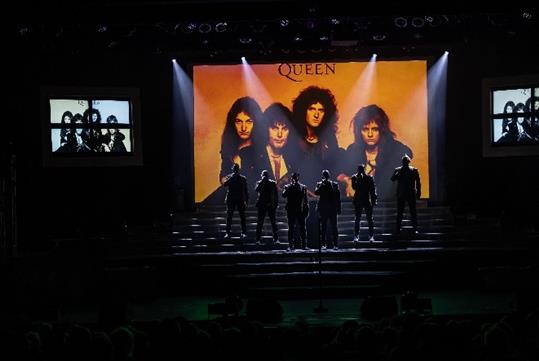 The Six Brothers performing on stage with an image of the iconic rock band Queen displayed on the LED screen as their backdrop.