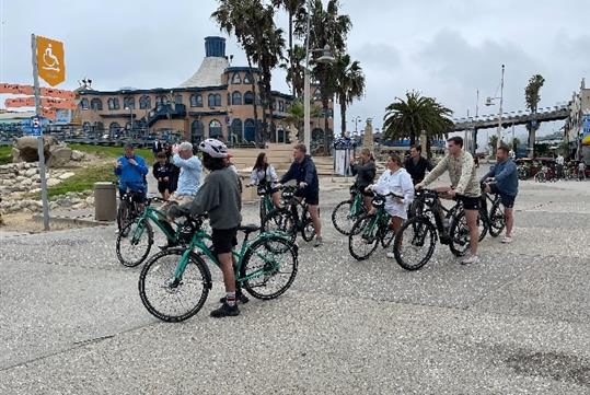 A group of tourists stopped on their bicycles and listening to their tour guide with a large building and palm trees in the background.