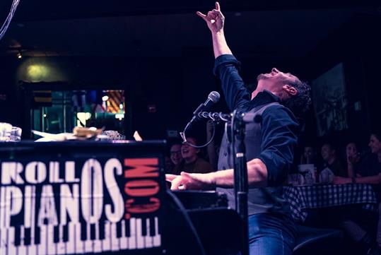 A man sitting and playing the piano with one of his hands in the air holding up a rock sign.