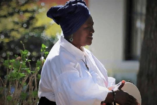 Master Gullah Geechee Truth-Teller ‘Sistah Patt’ wearing a blue turban and a white shirt while playing a tambourine outside.