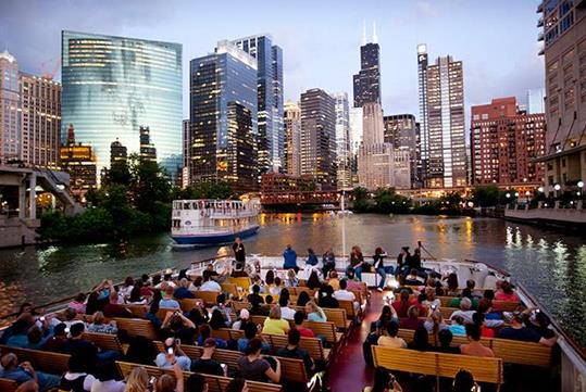 A large boat deck full of tourists sitting on benches listening to their tour guide at the front of the boat with the city of Chicago in the background at dusk.