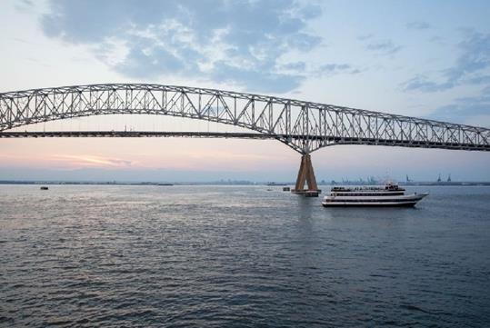 The Francis Scott Key Bridge in Baltimore, Maryland at sunset with a small cruise ship sailing past it.