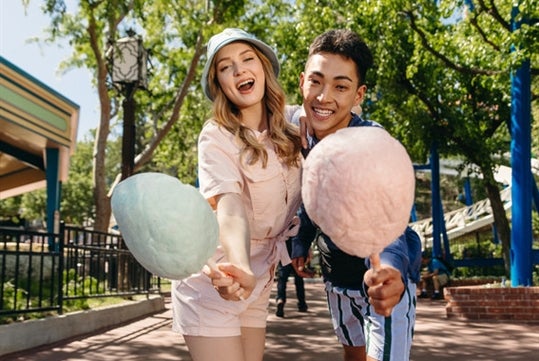 A couple smiles for picture while holding a pink cotton candy inside Six Flags America with trees behind them.