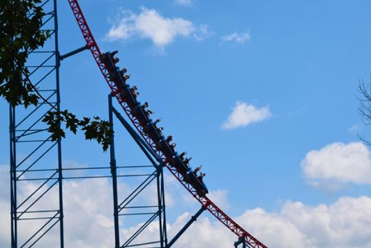 A roller coaster with a red track and a blue steel structure with riders seated with their hands raised in excitement and blue cloudy sky in the background.