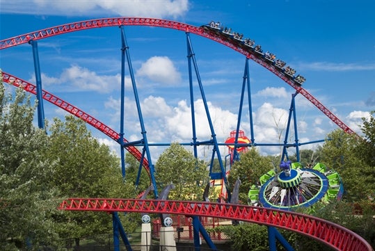 The SUPERMAN™ The Ride with a red track and blue supports moving against a clear blue sky with some clouds.