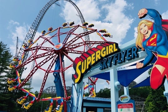 The "SUPERGIRL Sky Flyer" is a vibrant spinning pendulum attraction, featuring a predominantly red ride with yellow and blue accents, matching the Supergirl theme, while in the background, a large roller coaster with a massive red track loops into the sky on a clear, sunny day surrounded by lush green trees at Six Flags St. Louis.