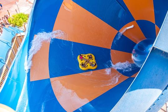 Aerial view looking down on a group of four people sitting in a yellow raft on a blue and orange checkered waterslide.