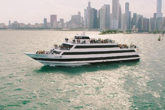 A cruise boat with all it's desk full of people having a good time on a sunny day with the city of Chicago behind them.