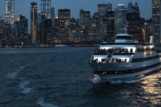 A cruise boat with people standing on it's deck at night with the city of New York in the background.