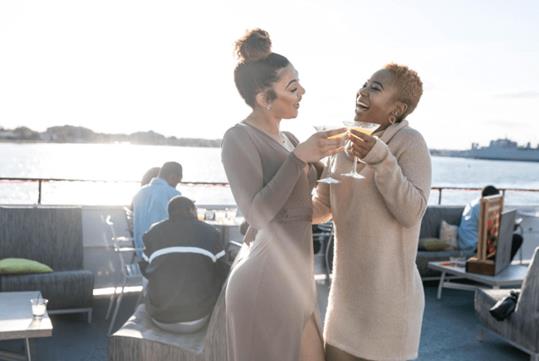 Two women in beige dressed standing together and laughing while cheering their drinks together on the Spirit of Norfolk Signature Brunch Cruise.