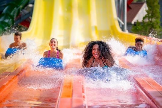 Four guests splashing into water at the bottom of a yellow and orange waterslide at Splish Splash Water Park.