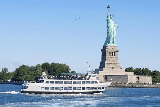 A white cruise boat with tourists standing on it's top deck sailing past the Statue of Liberty on a sunny day on the Statue of Liberty & Ellis Island with Round-trip Ferry.