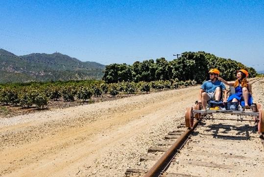 A man and woman wearing orange helmets while riding the Sunburst Railbikes and enjoying nature on a sunny day near Los Angeles.