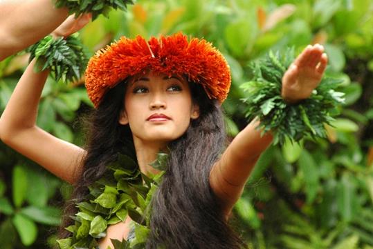 A beautiful hula dancer wearing an orange headdress with her hands up wearing bracelets and at The Feast at Mokapu Luau in Maui, Hawaii, USA