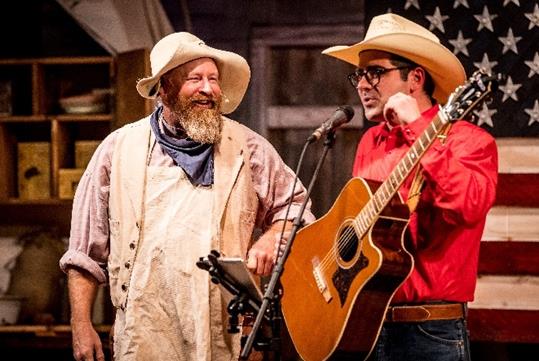 A man in a beige western costume next to a man in a red shirt and cream cowboy hat with a guitar performing on stage at The Great American Chuckwagon Dinner Show.