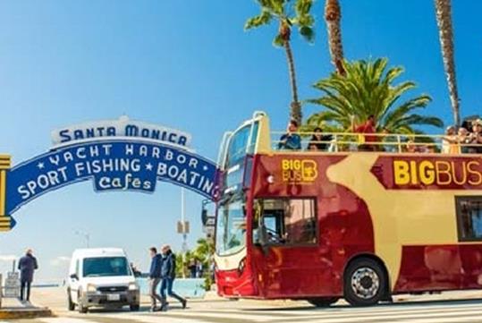 A Big Bus tour bus with tourist filling the top level driving by the for the Santa Monica Yacht Harbor on a sunny day.