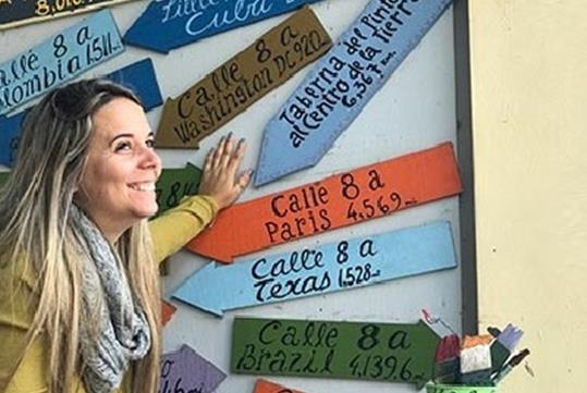A blonde woman in a yellow shirt smiling with her hand on a wall of multicolored arrow signs next to her on the Little Havana Adventure.