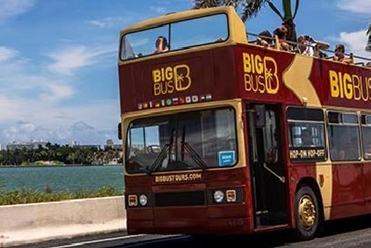 A maroon double decker Big Bus with the top level full of tourists on a sunny day with water and trees in the background.