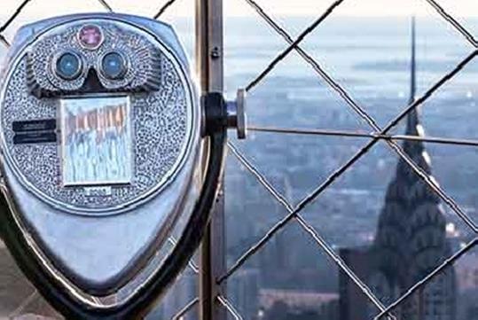 A coin operated telescope in front of a fence with the city of New York in the background at the Empire State Building.