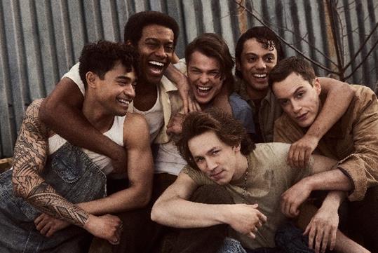 The cast of The Outsiders in costume huddled together laughing with a tin wall behind them.