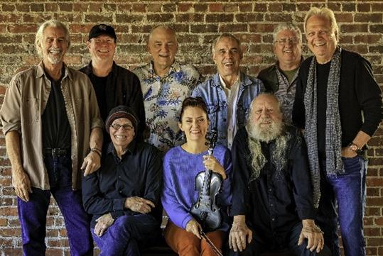The smiling members of The Ozark Mountain Daredevils posed with six standing and three sitting in front of a brick wall.