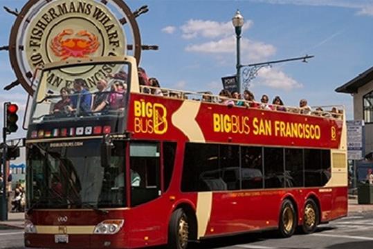A Big Bus San Francisco tour bus with the top deck full of people driving by Fishermans Wharf on a sunny day.