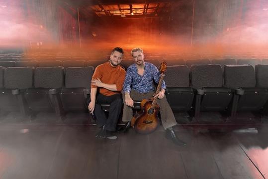 A man in a dark orange shirt sitting next to a man in a blue shirt with a guitar at The Sound of Simon & Garfunkel.