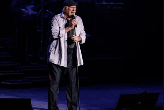 Terry Bradshaw on stage, smiling and singing into a microphone, dressed in a casual outfit with a striped shirt and cap, while a band is visible in the background.