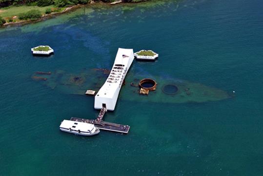 Aerial view looking down on the USS Arizona Memorial with deep blue green water around it on a sunny day on Oahu.
