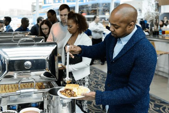 A man in a blue sweater ladling food onto his plate at the end of a buffet line with other guests in line behind him on the Washington DC Premier Brunch Cruise.