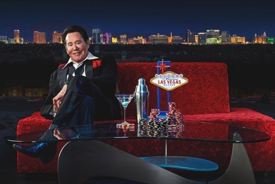 Wayne Newton sitting on a red sofa with Las Vegas as the background.