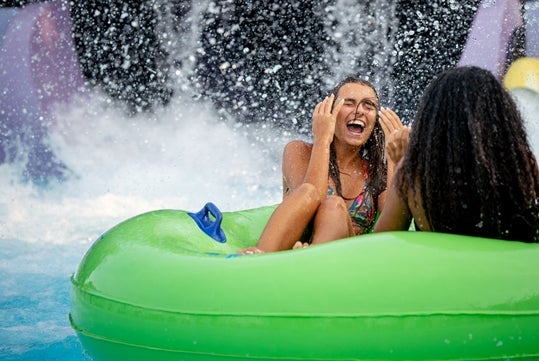 Two women sit facing each other on a green inflatable tube, laughing with joy as water splashed behind them