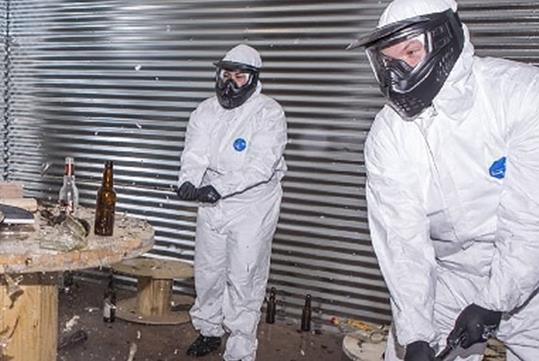 Two men in white suits wearing protective face gear and smashing glass bottle at Wreck Room A Destruction Experience.