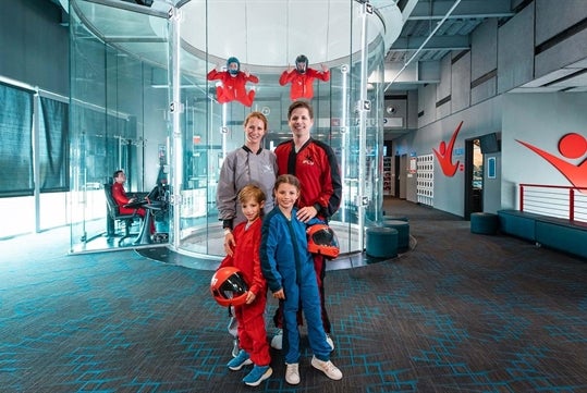 A family in jump suits standing in front of a flight tunnel with two instructors floating in the air behind at iFly.