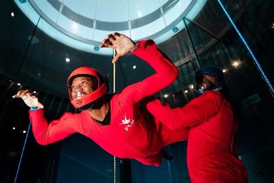 Two men in red jump suits in a flight tunnel, one of them flo aating in the air and the other holding onto his suit guiding him.