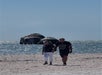 A couple walks on a sandy beach while old, dome-shaped structures sit partially submerged in the ocean in the background.