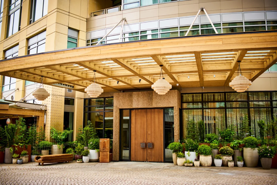 The entrance of a modern building featuring large double wooden doors, hanging lights, a wooden pergola, and potted plants arranged along the entryway.