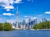 Toronto skyline with the CN Tower and modern skyscrapers viewed from across the water, with trees and boats in the foreground under a blue sky.