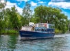 A blue and white passenger boat travels on a calm river, with green trees and a partly cloudy sky in the background.