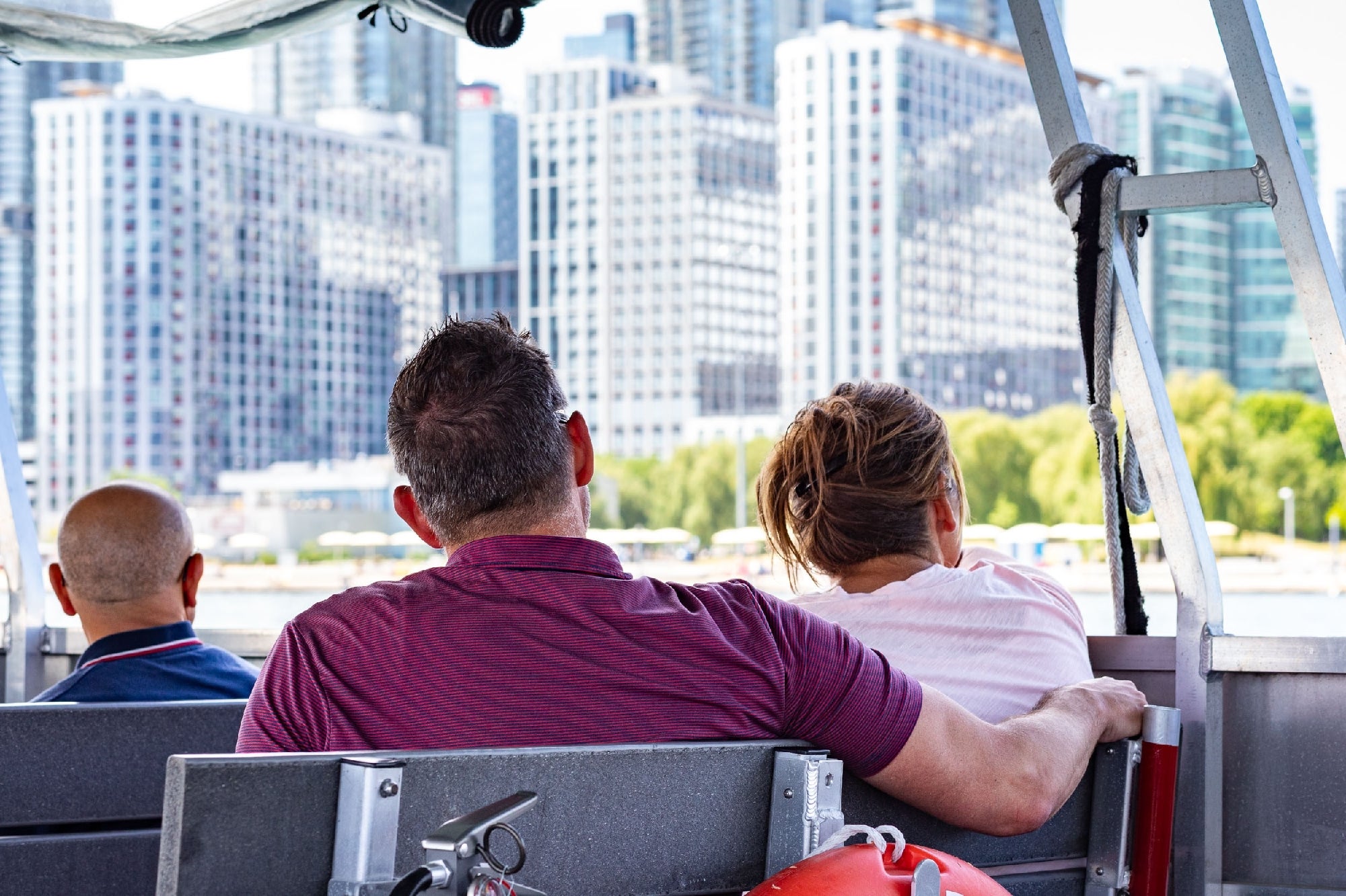 Three people sit on a boat facing a city skyline with tall modern buildings in the background.