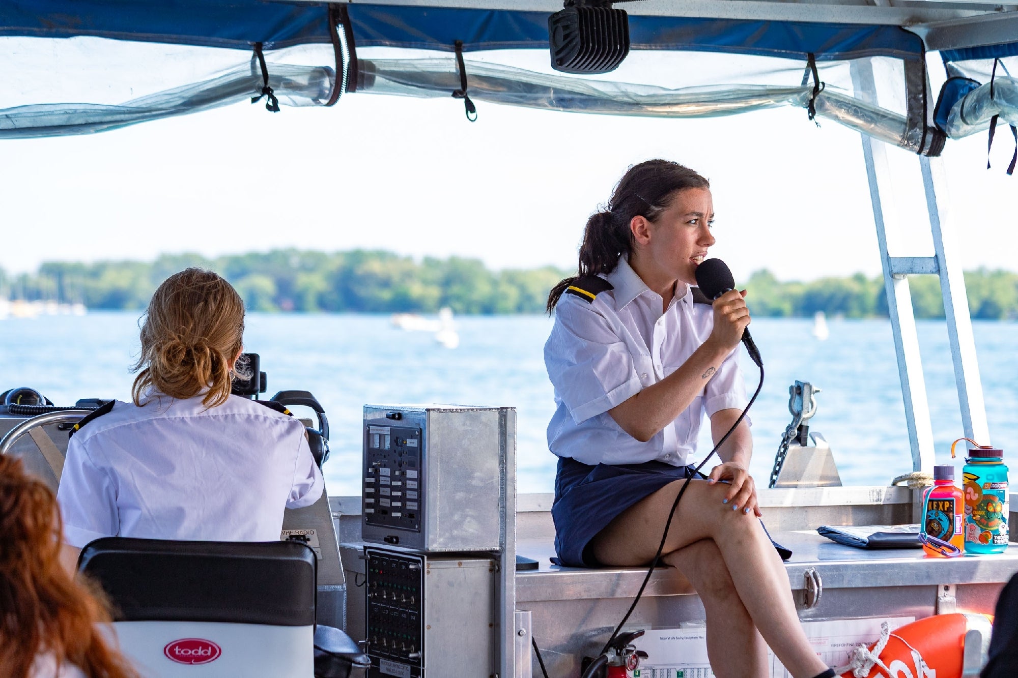 Two women in uniform operate a boat; one steers while the other speaks into a microphone. Trees and water are visible in the background.