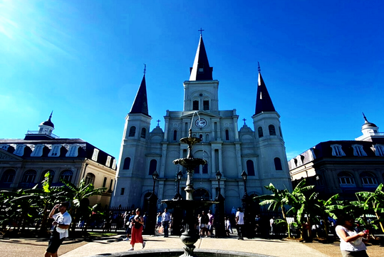 People gather in front of a historic cathedral with three spires and a central clock, under a clear blue sky, with a fountain and palm trees in the foreground.