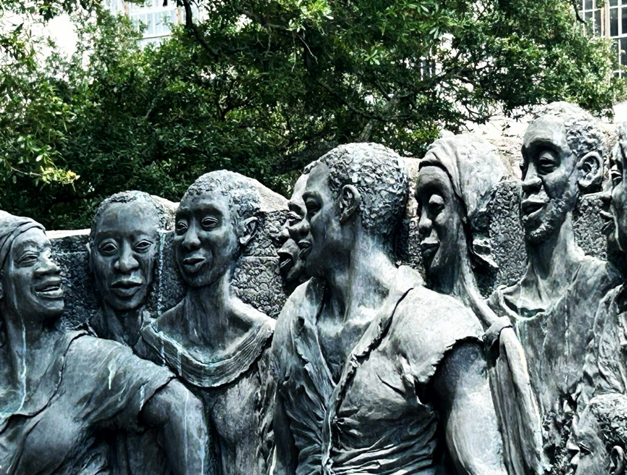 A bronze sculpture depicting a group of people standing close together, smiling and interacting, with trees and buildings in the background.